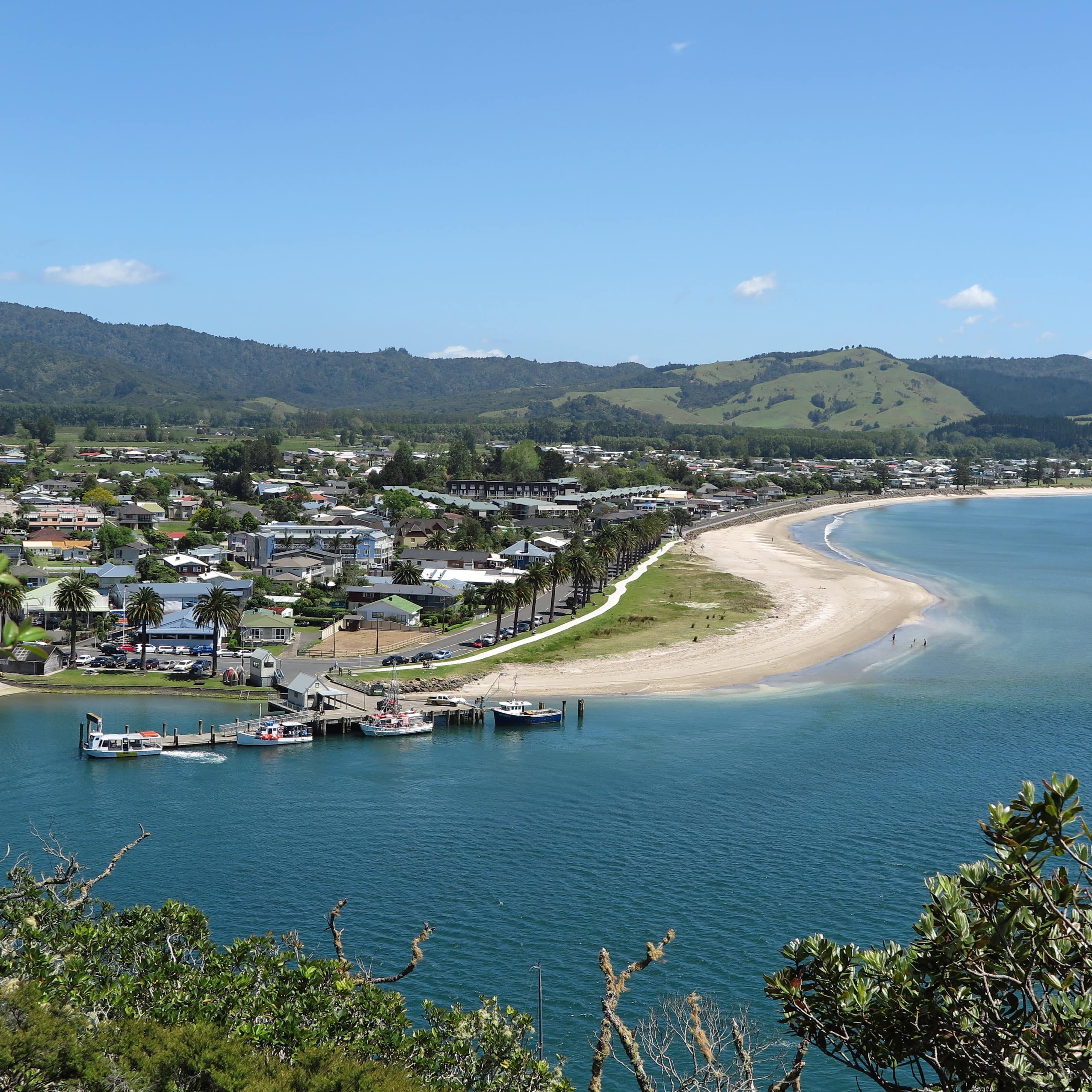 Seaside town, blue waters, and coastline of Whangamata Beach with dark green grass covered hills in the background