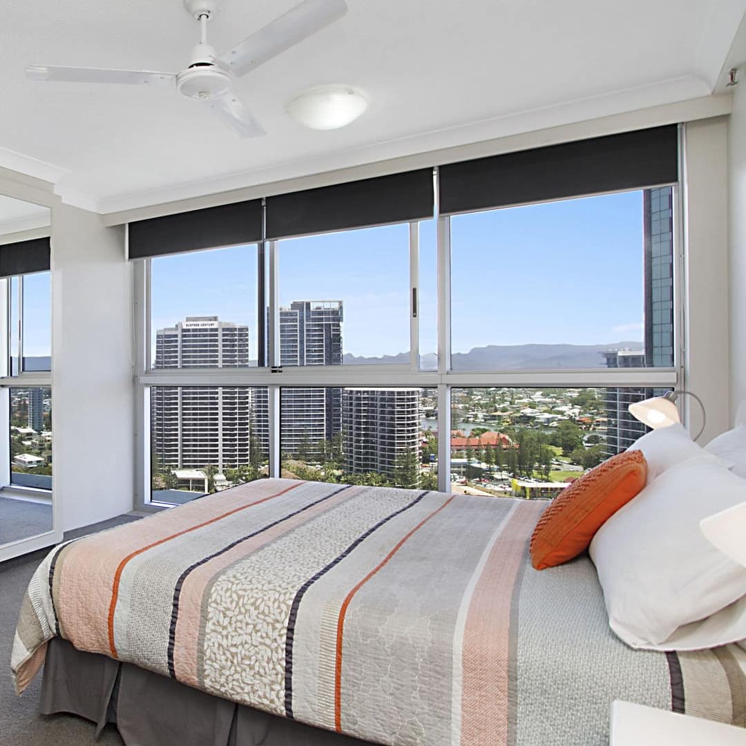 Bedroom with grey carpet, a king size bed, orange and grey bedding, mirrored closet doors, and tall windows.