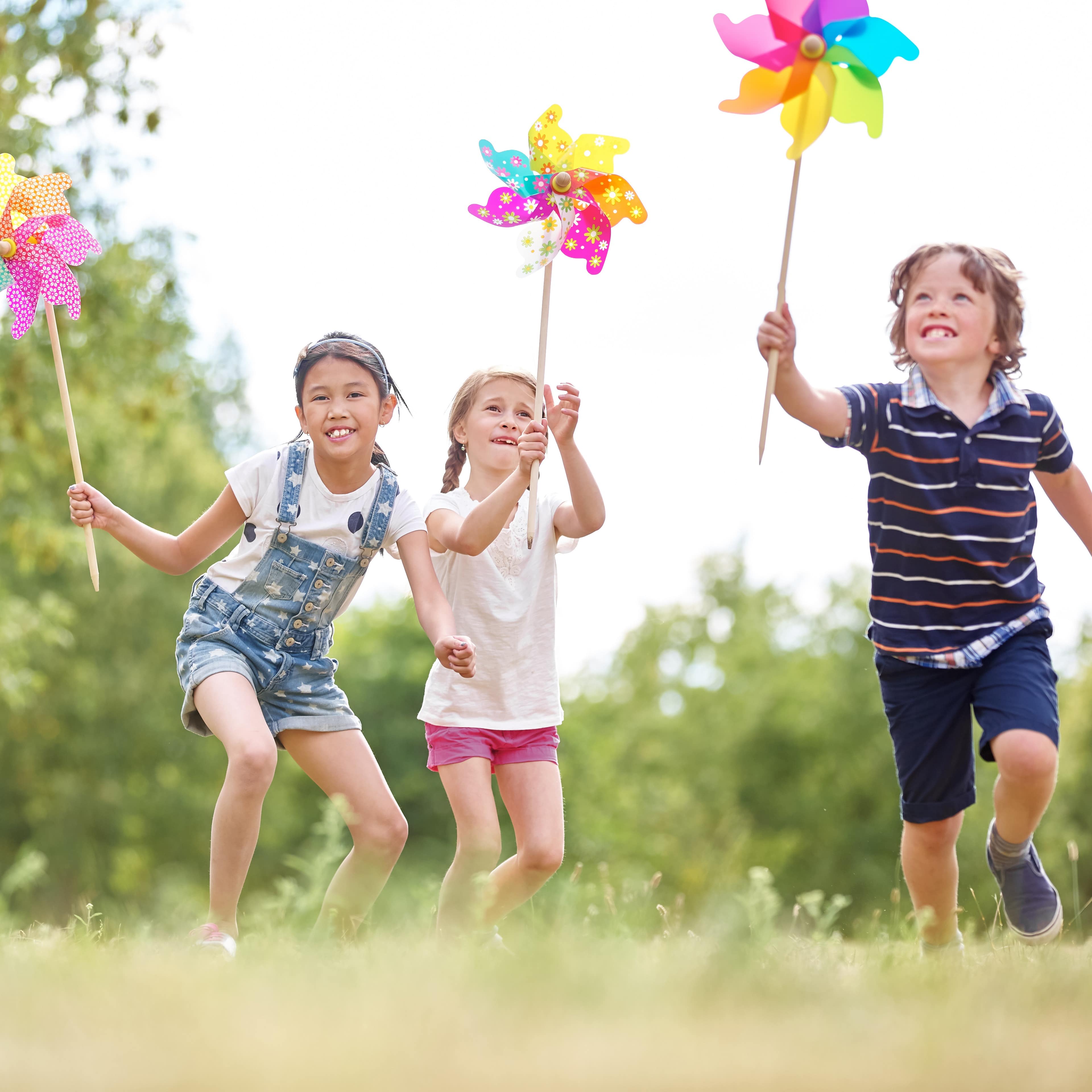 Three children running with pinwheels in a green open space at the park