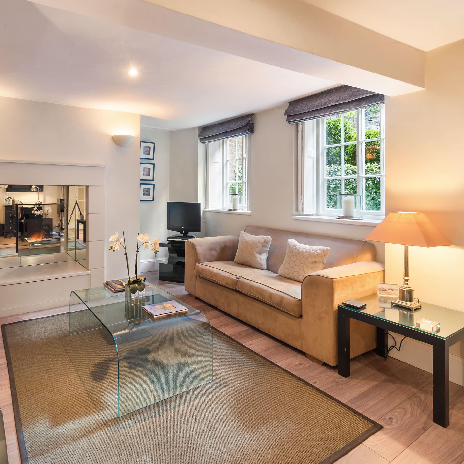 Modern living room with hardwood floors, beige suede sofas, acrylic coffee table, and mirror-framed gas fireplace.