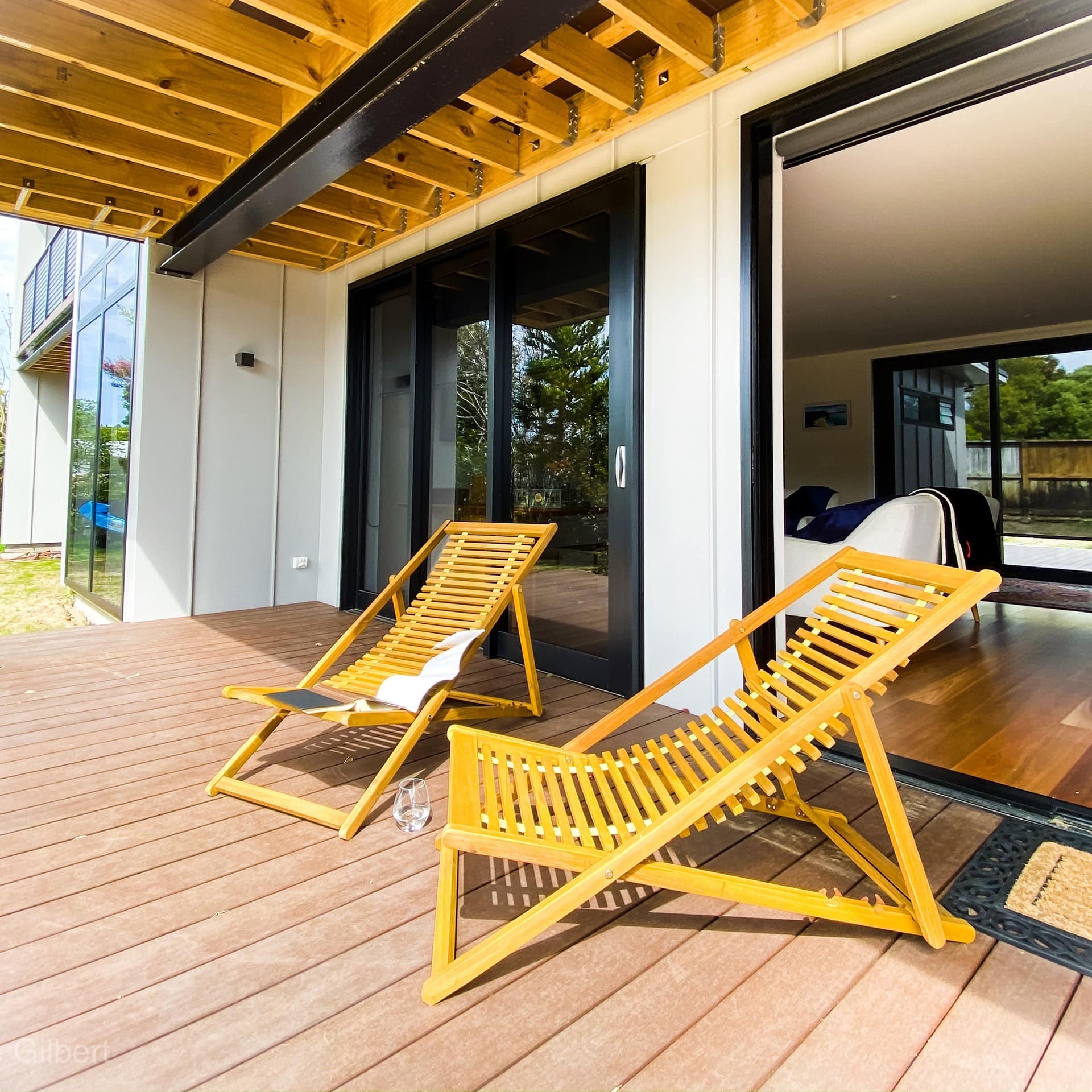 Two wooden beach chairs on the deck of a contemporary beach house with a view of trees and the blue sky through the windows