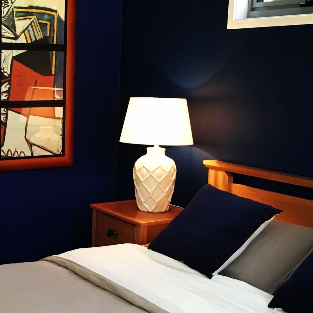 Bedroom with navy blue walls, wood-framed bed with grey and white bedding, wood bedside table, modern art.