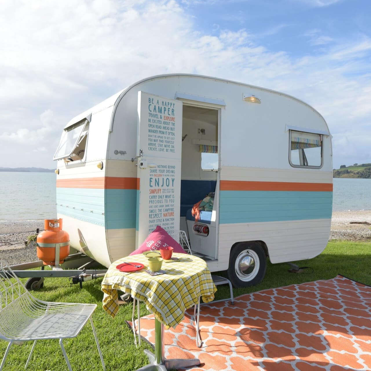 A colourful caravan on the shore of Maraetai beach with a small table setting and picnic mat in front