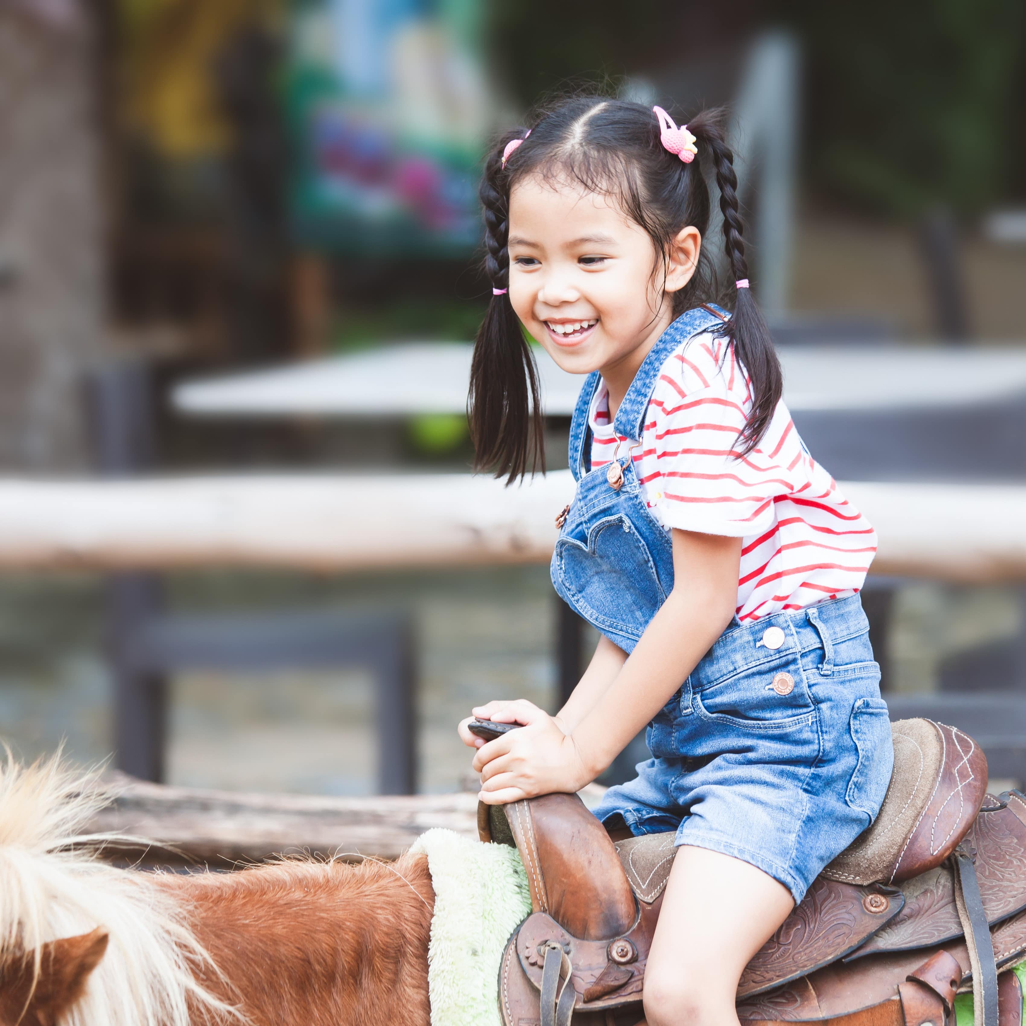 Young girl with pigtails and overalls riding a pony on a farm