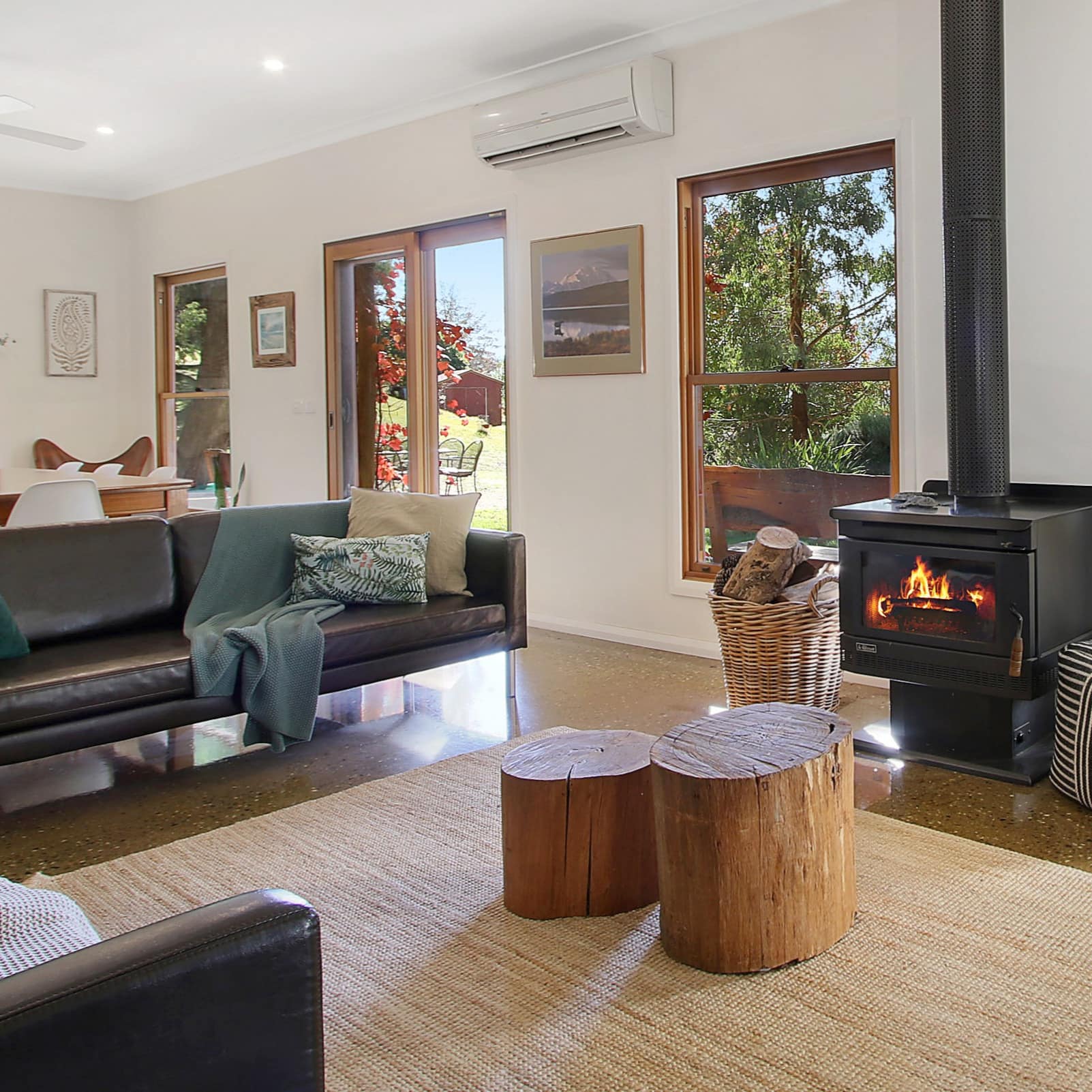 Open living and dining room with stone floor, black leather sofas, wood-burning stove, tree stump seats, and a woven rug.