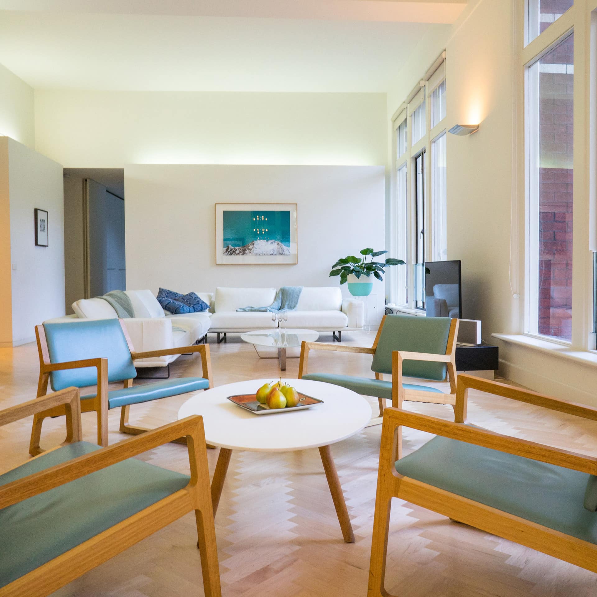 Open living room with chevron wood floors, white walls, floor to ceiling windows, and modern furnishings.