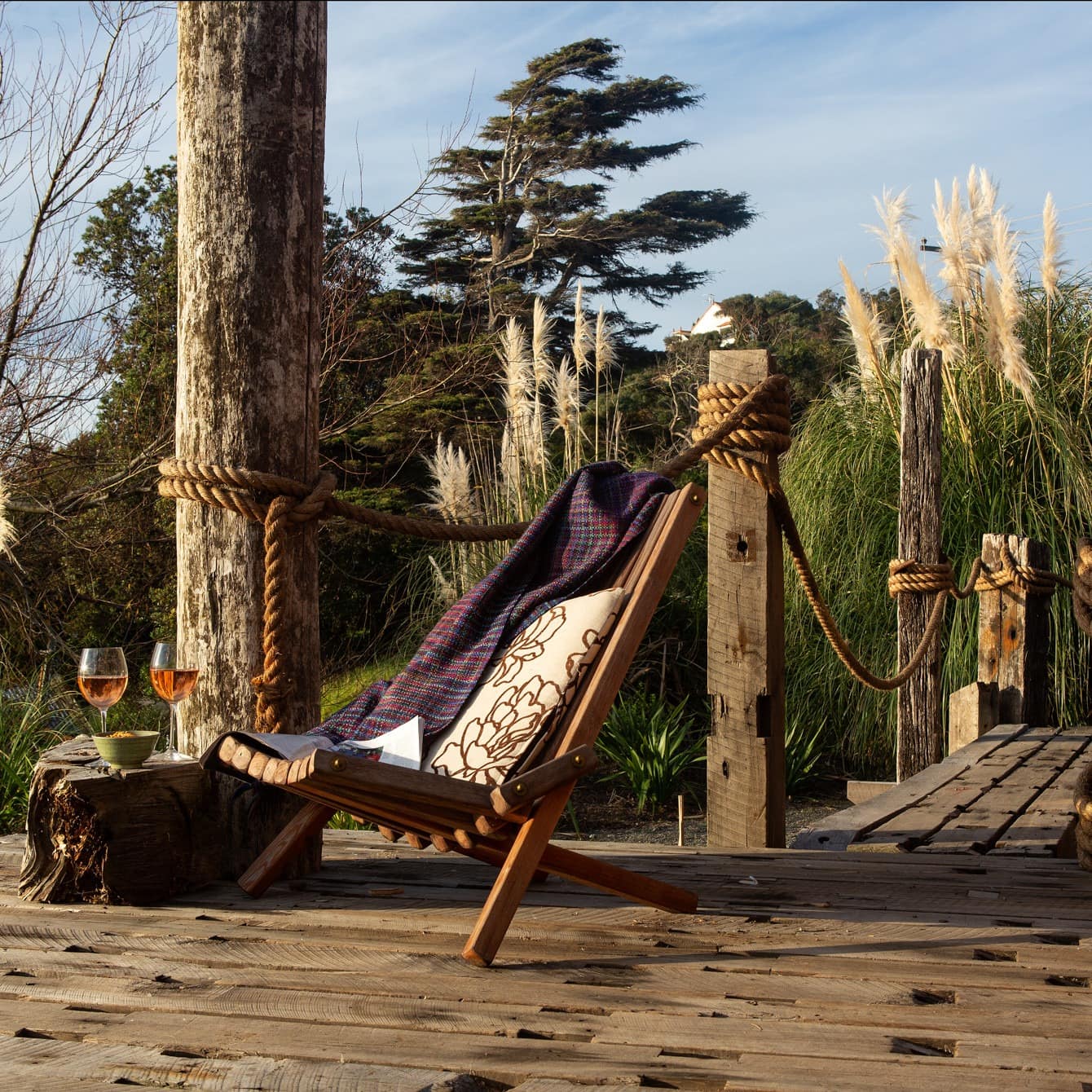 A cozy reclined chair with two wine glasses sitting on a wooden deck with greenery in the background