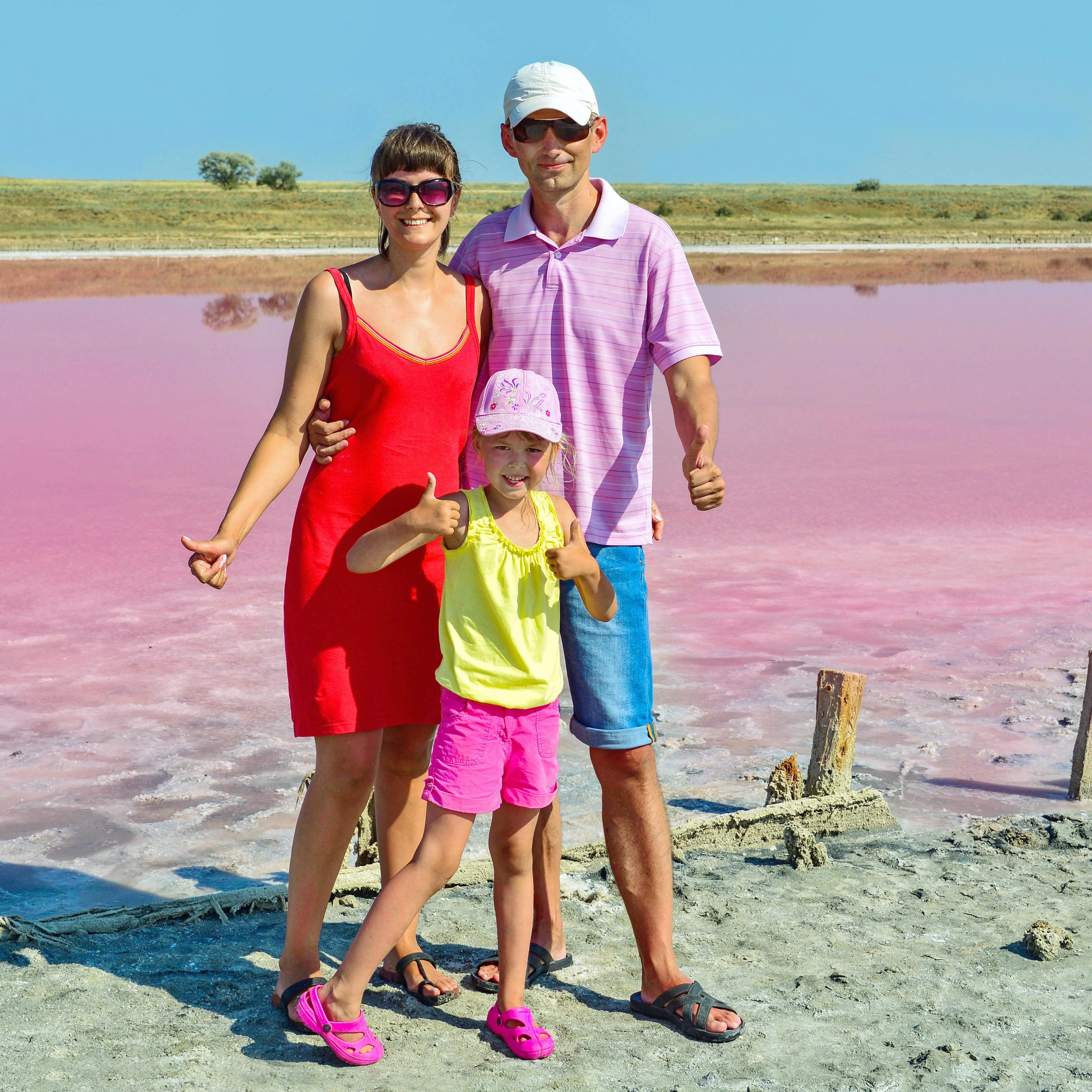 Family of three in bright colors with thumbs up as they stand in front of pink salt lake with gold hills in background