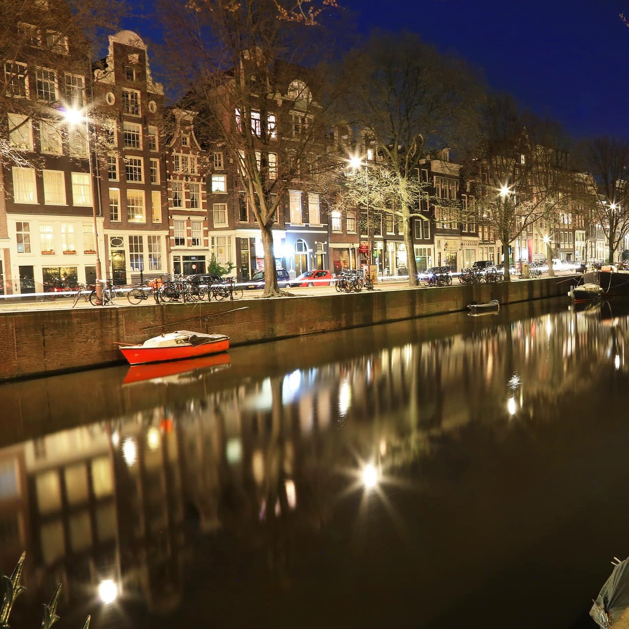 An Amsterdam canal with canal houses, trees, streetlights, boats and bicycles parked on the sidewalk